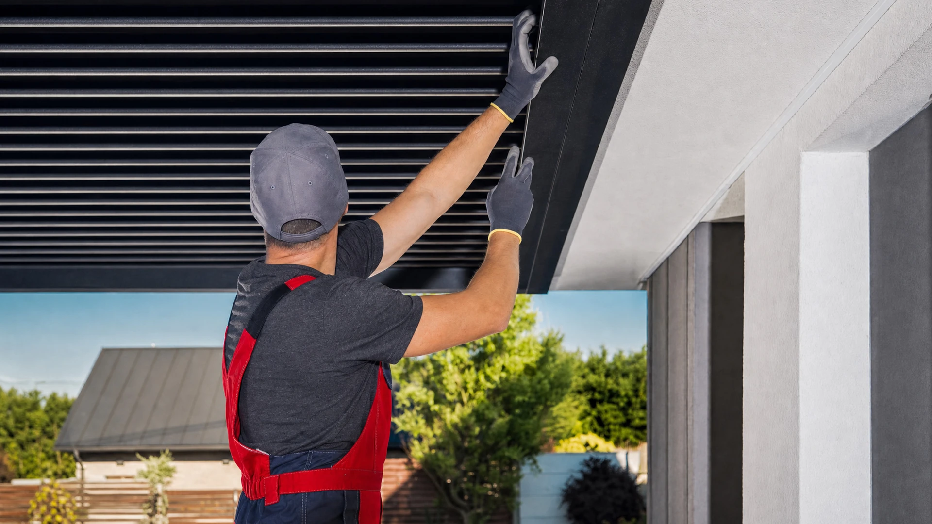 Technicien menuisier en tenue professionnelle ajustant les rails et ressorts d'une porte de garage sectionnelle aluminium gris anthracite sur la façade d'une maison résidentielle, avec un tournevis en main et des outils disposés à proximité.