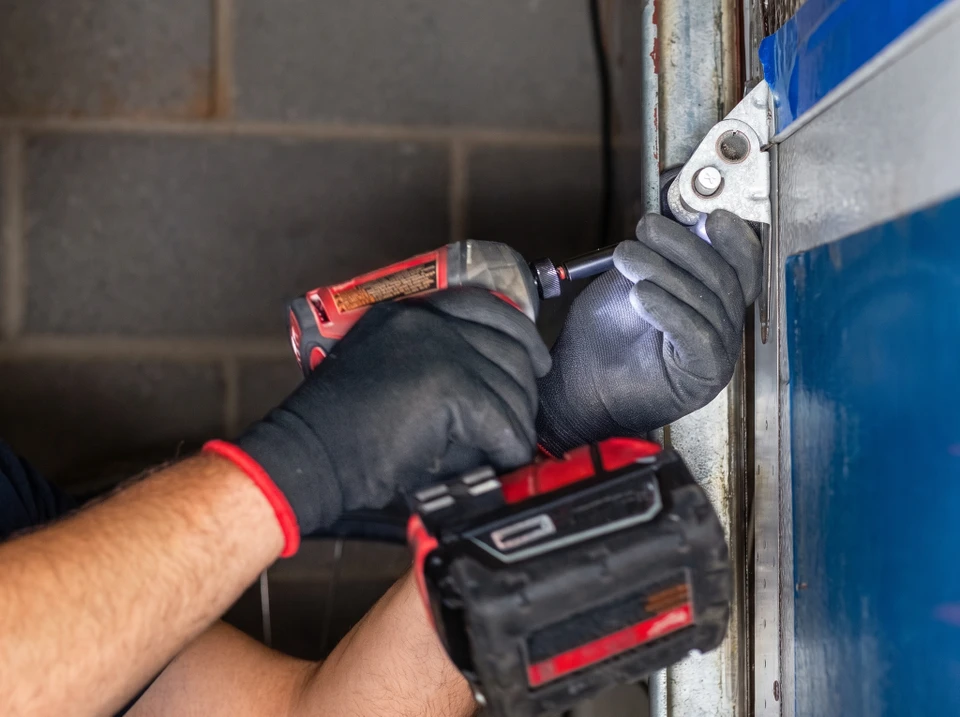 Technicien menuisier en combinaison de travail, agenouillé ou penché, effectuant le diagnostic et la réparation d'une porte de garage sectionnelle blanche immobilisée dans le garage d'une maison résidentielle, avec ses outils à proximité.