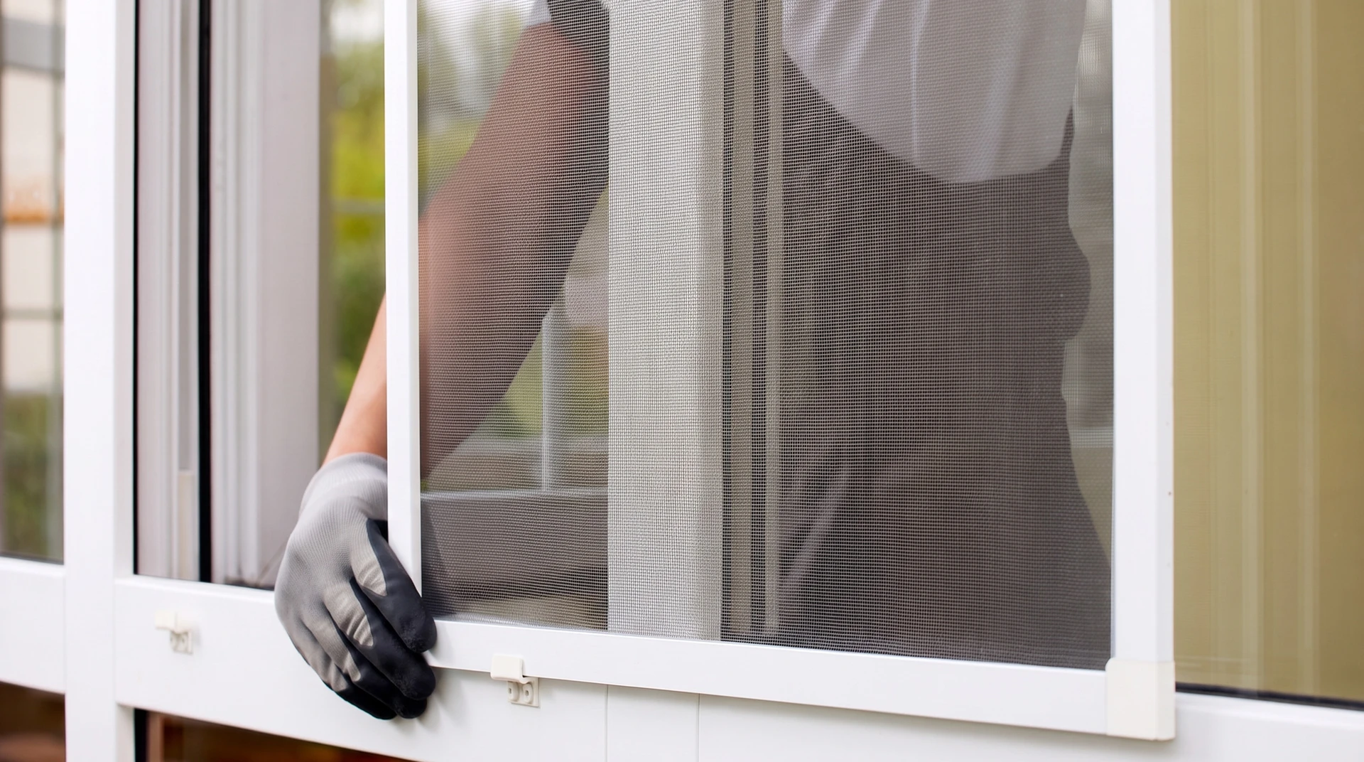 Un ouvrier portant une casquette noire et un polo bleu installe une moustiquaire sur une fenêtre à battants depuis l'intérieur d'un appartement en hauteur