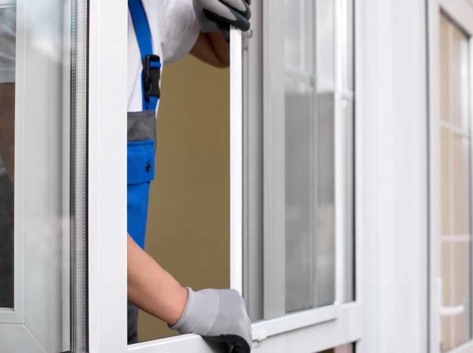 Artisan menuisier mesurant précisément l'embrasure d'une porte d'entrée vitrée avec un mètre ruban, en préparation de la pose d'une moustiquaire sur mesure, devant une maison individuelle en journée.