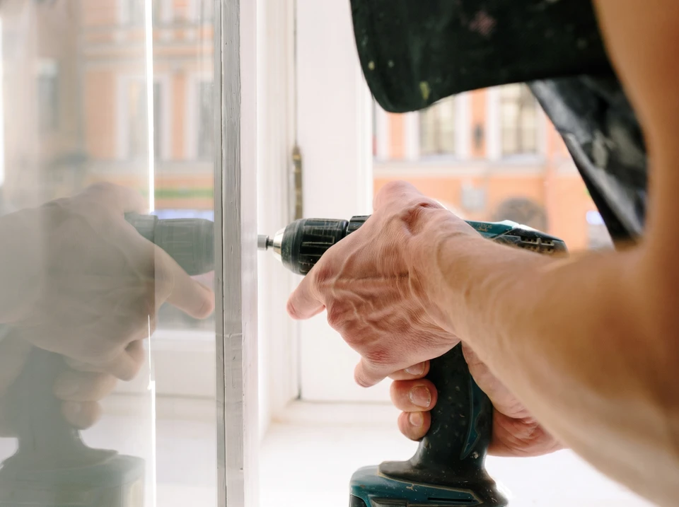 Artisan menuisier barbu en tenue professionnelle discutant face à face avec un client propriétaire devant une fenêtre extérieure en bois clair, l'artisan pointant du doigt un détail de la menuiserie tandis que le client l'écoute attentivement, scène dans l'extérieur d'une maison d'habitation en journée.