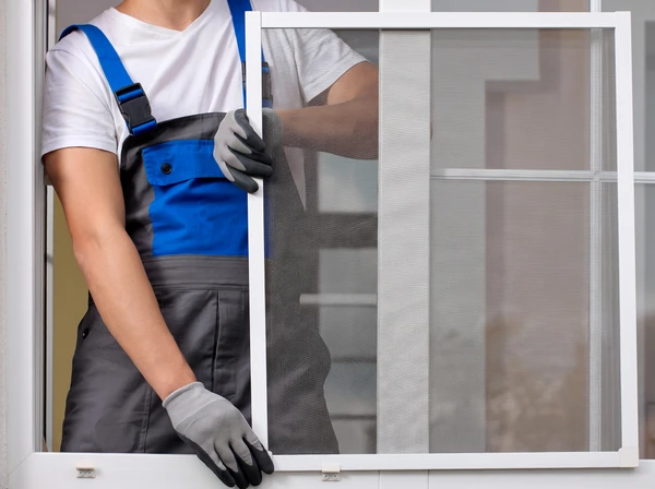 Ouvrier en salopette bleue et casquette installant une fenêtre coulissante sur façade extérieure d'habitation